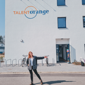 Una joven pelirroja frente al edificio de la escuela de idiomas TalentOrange. El edificio es blanco y lleva las letras TalentOrange en naranja y azul. La mujer señala invitadoramente hacia la puerta abierta.